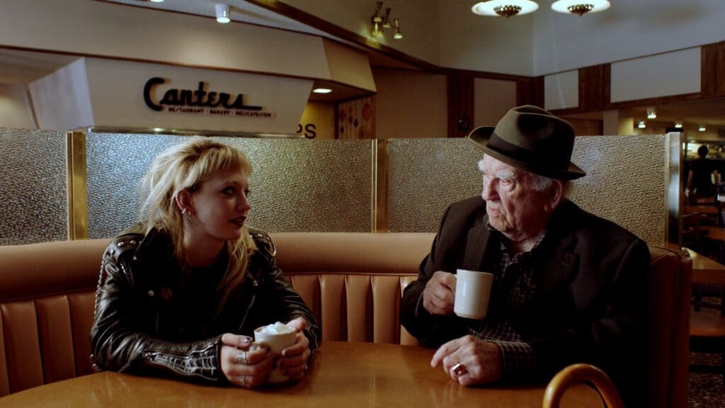 A yong woman in alternative clothing sits with an older man in a cafe. They are both holding cups of coffee. 