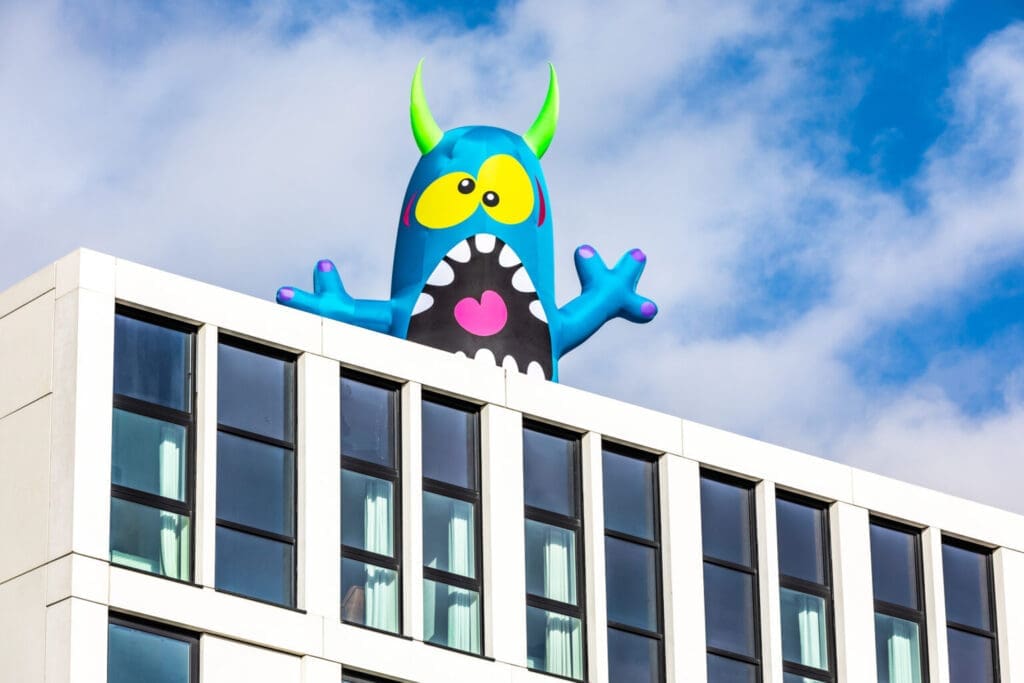 A blue inflatable monster sits above a white building. The monster has yellow eyes and green horns.