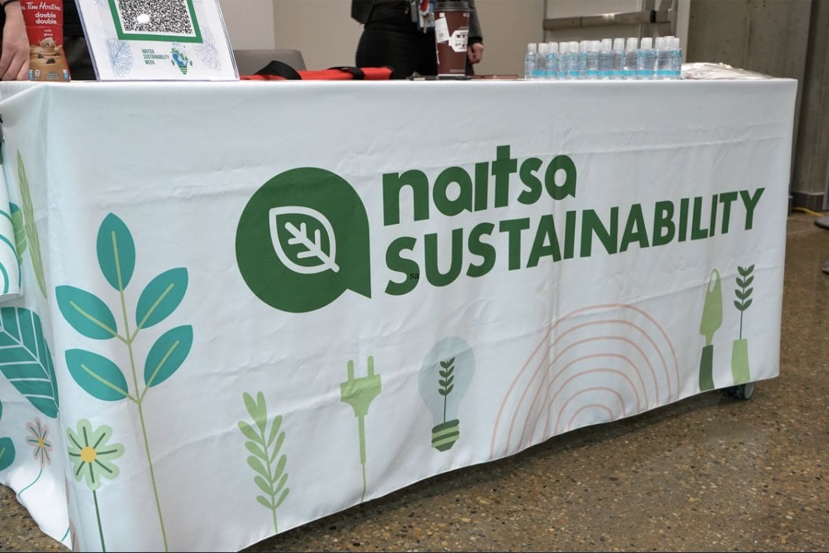 naitsa sustainability week A white tablecloth covers a table. It says "naitsa sustainability" in green letters. there are green and light blue leaves on the bottom.