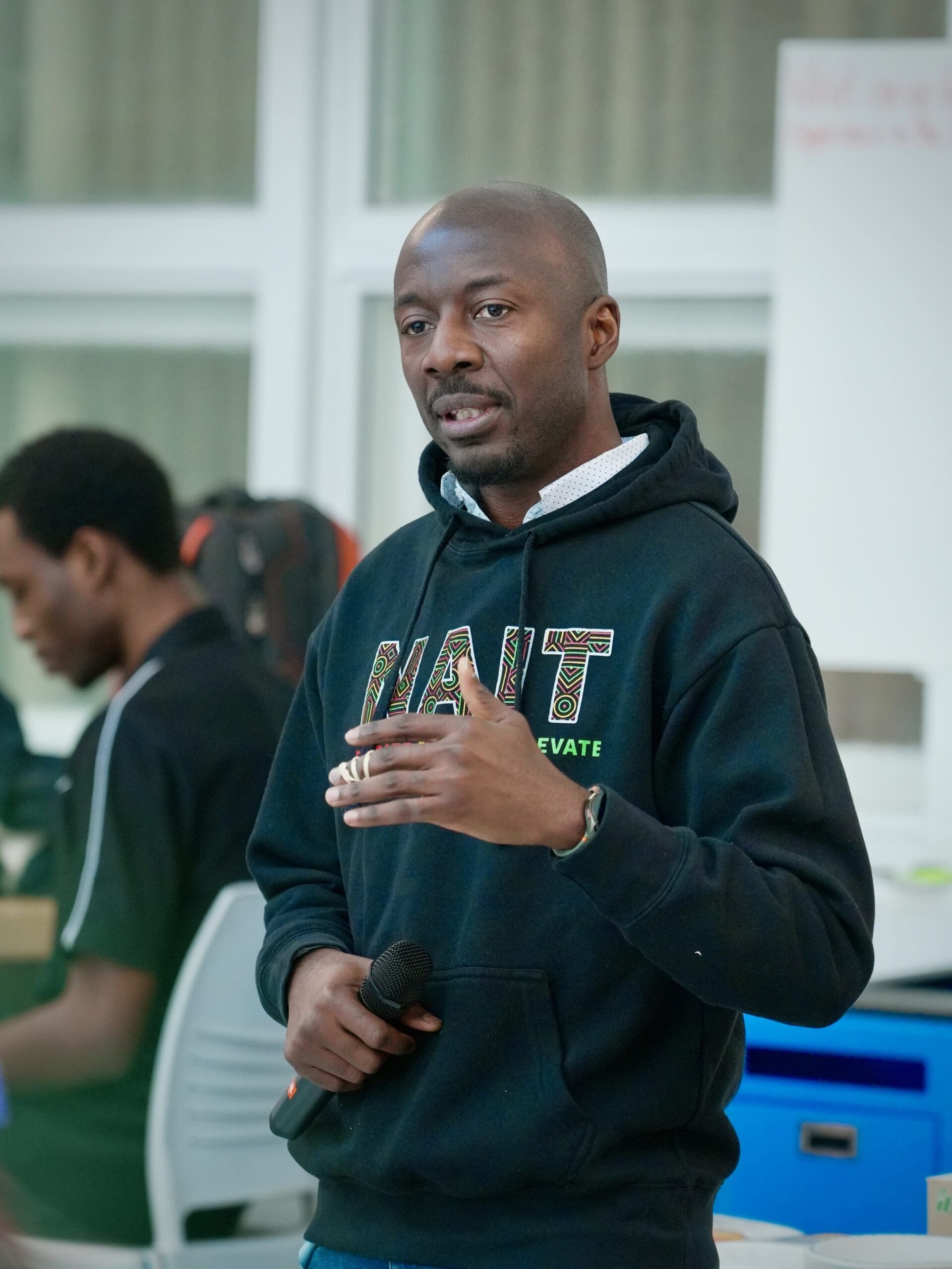 A bald Black man wearing a NAIT hoodie speaks to a crowd. 