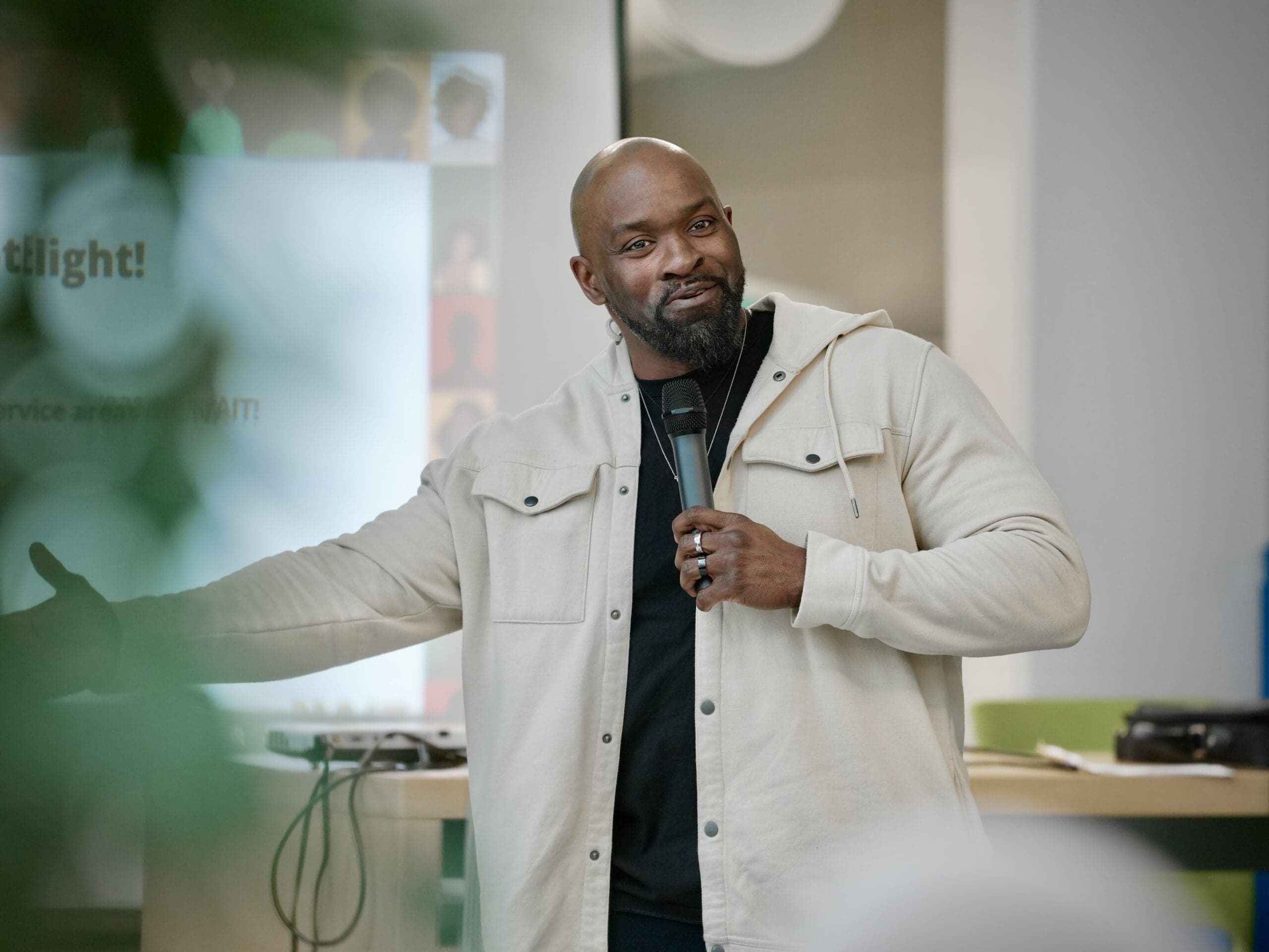 A tall Black man wearing a beige sweater speaks into a microphone.