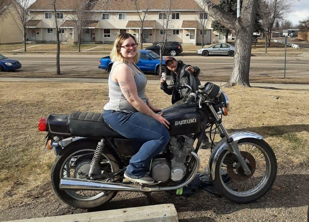 Samantha poses on a Suzuki motorbike in a residential area. In the background, a male family member wearing leather points to the camera and poses.