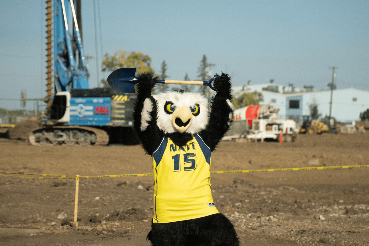 ASC groundbreaking NAIT’s Ook mascot raises a shovel above its head at a construction site, wearing a yellow and blue NAIT No. 15 jersey. An excavator and construction equipment are visible in the background.