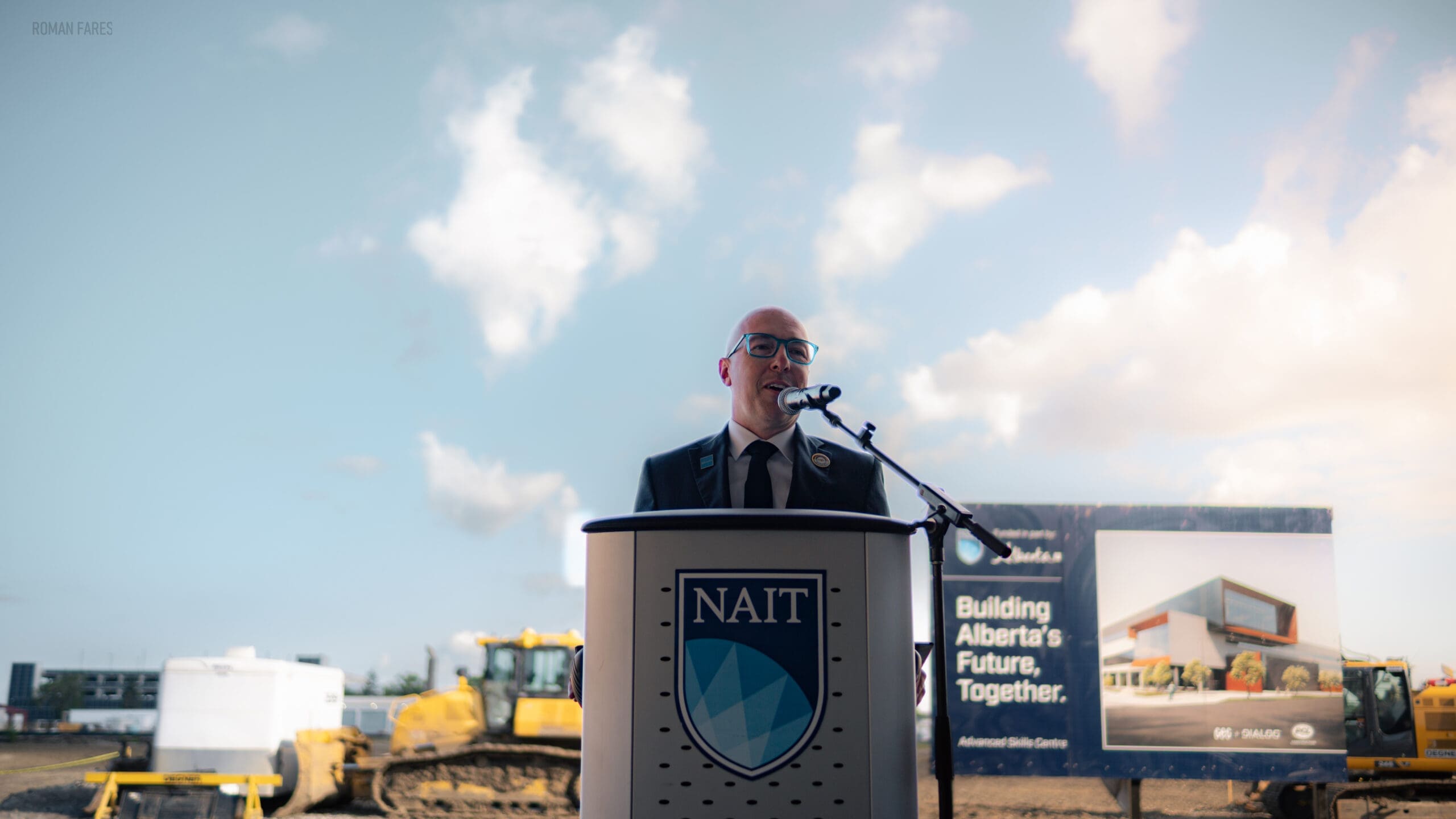 Edmonton mayor Andrew Knack speaks at a podium during the Advanced Skills Centre groundbreaking ceremony. Construction equipment and a rendering of the future building are visible behind him.