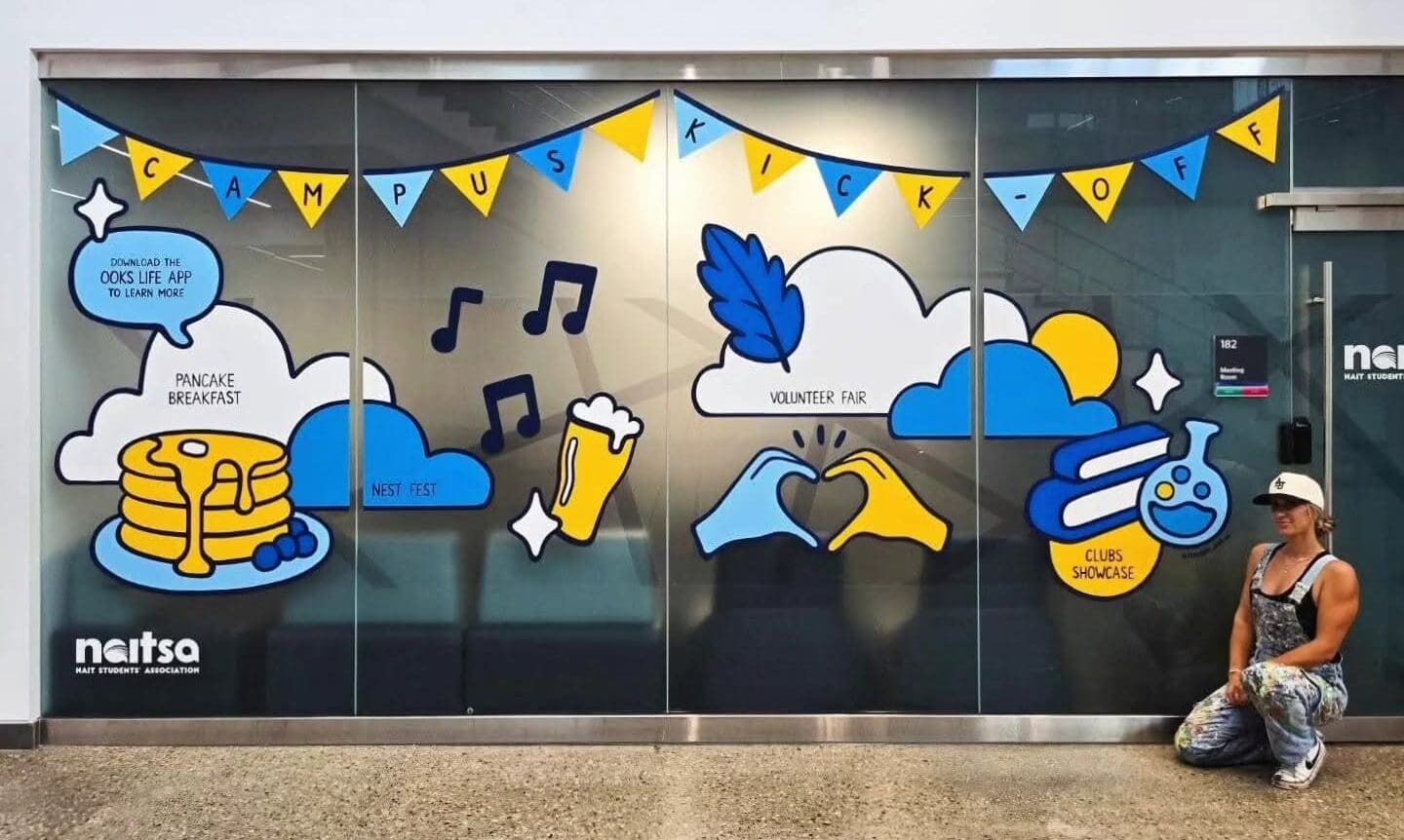 Wide shot of NAIT's newest vivid window art, behind Alixandra posing beside it.