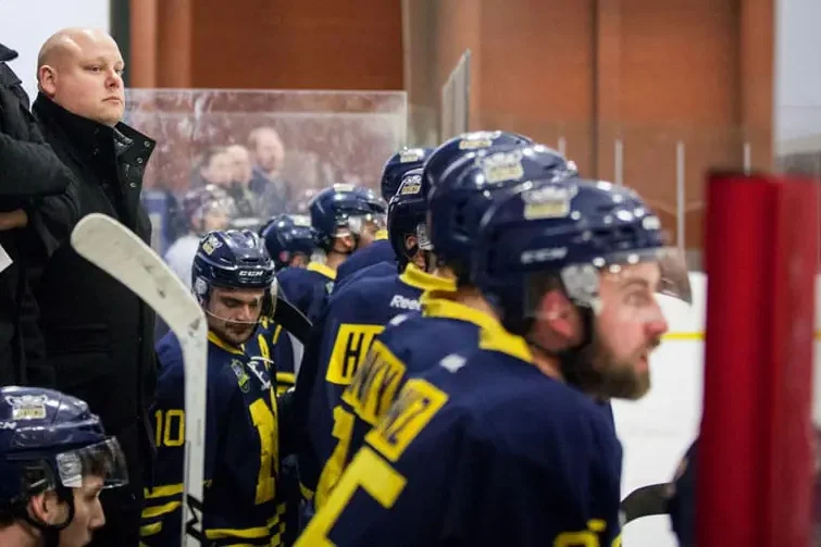 Tim Fragle in the upper back corner of the background, looking down to his team in the foreground during a hockey match.