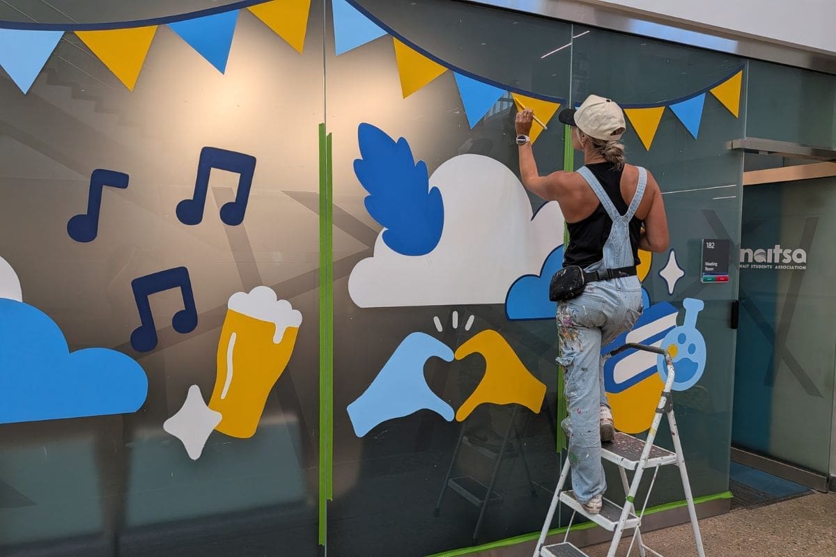 Photo of Alixandra Jade in the process of painting the NAIT room mural, standing on a ladder while painting a banner piece.