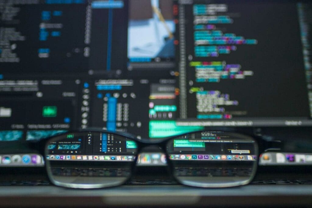 Cover image. Photograph of cluttered computers screens all displaying blurry coding software and code, through the lens of glasses set Infront of the computer, a small portion of the computer screens become visible through the lenses of the glasses.