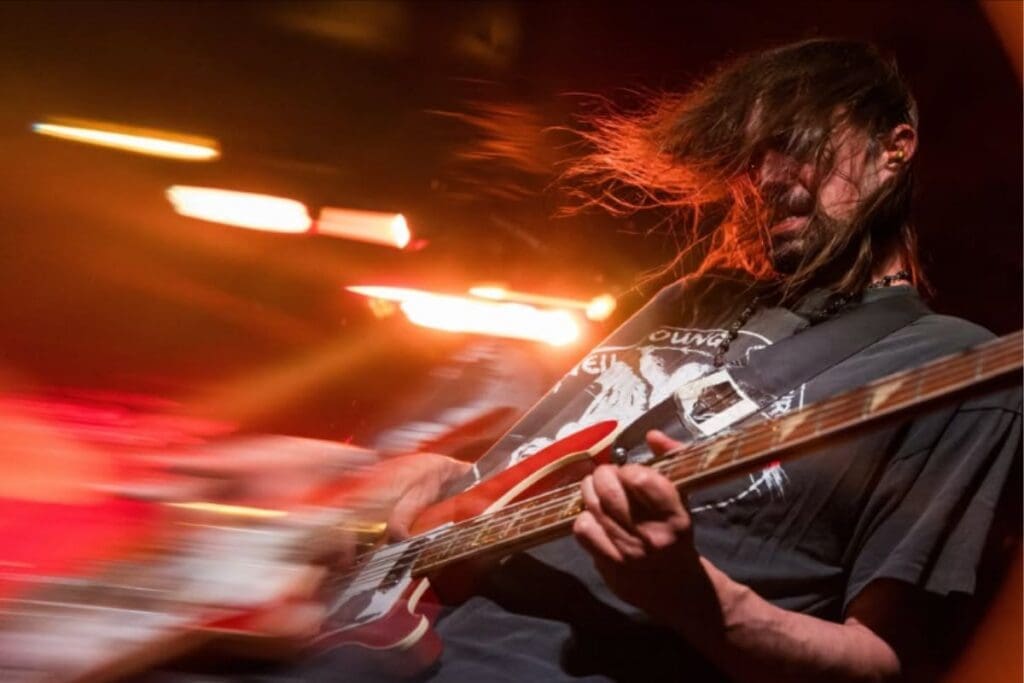 Feature image. Closeup Photograph of a person playing a guitar within an event, the background is blurry and gives a blurry stop motion effect.